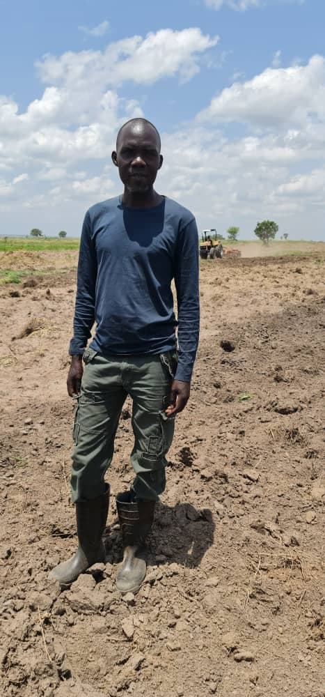 Rama Omonya standing on a freshly ploughed field with a tractor working behind him